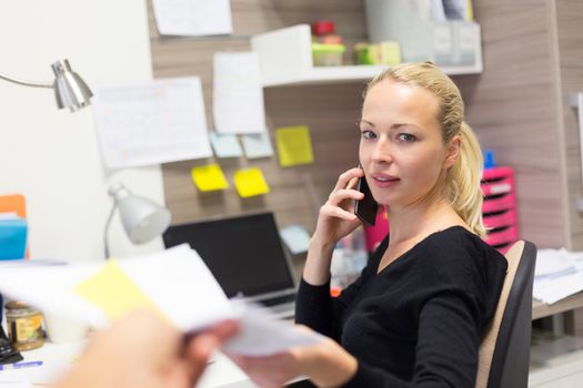 Business and entrepreneurship consept. Beautiful blonde business woman talking on mobile phone in colorful modern creative working environment accepting papers. Female multi tasking.