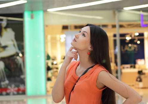 Pensive beautiful Asian girl in orange blouse in a large shopping center.