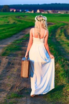 Blonde girl wearing white long dress with retro styled suitcase on countryside