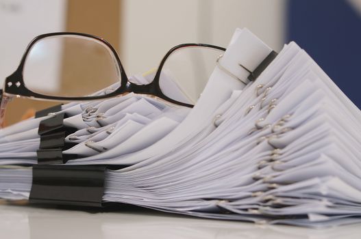 Glasses placed on finished document, stacks of paper files with clips on desk for report in the office at morning, Business offices concept.                              
