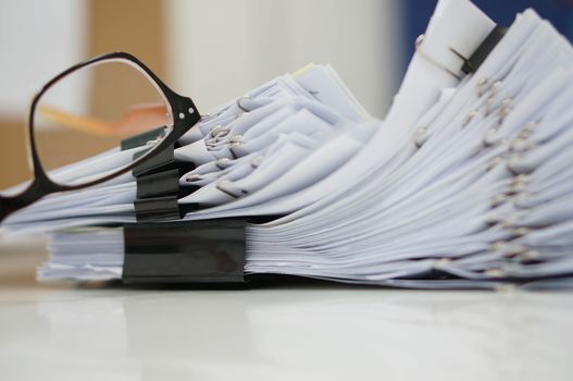 Glasses placed on finished document, stacks of paper files with clips on desk for report in the office at morning, Business offices concept.                     