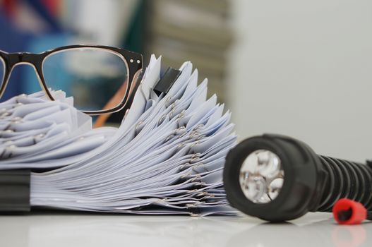 Unfinished document, stacks of paper files with clips on desk for report, glasses and flashlight in the office at evening, Business offices concept on workplace background.                                  