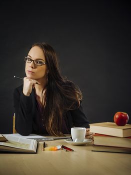 Photo of a teacher or business woman in her 30's sitting at a desk in front of a large blackboard.
