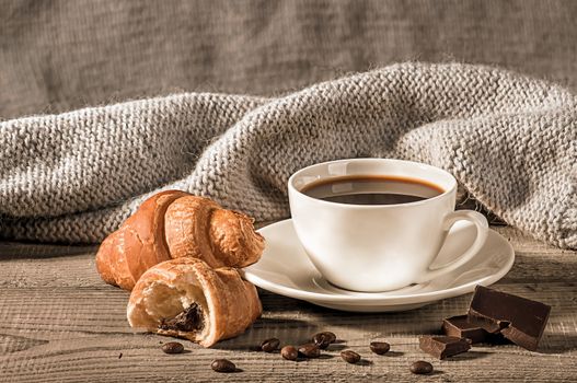 Coffee with croissants on the background of a woolen scarf. Grains of coffee next to the cup. Several pieces of dark chocolate next to each other.