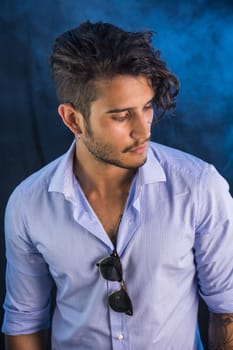 Portrait of brunette young man in light blue shirt and jeans, standing in studio shot against dark background.