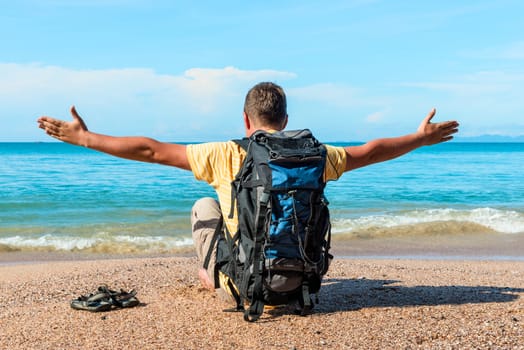 a tourist with a backpack with arms spread out to the sides sits on the sand near the sea