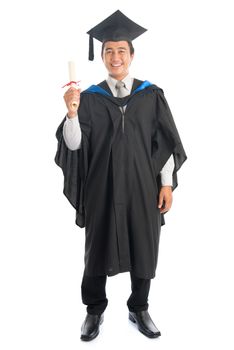 Full body male university student in graduation gown holding paper certificate, standing isolated on white background. Attractive Southeast Asian model.