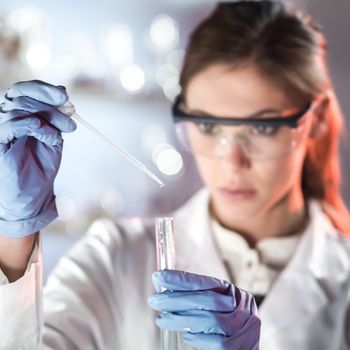 Life scientists researching in laboratory. Focused female life science professional pipetting solution into the glass cuvette. Lens focus on researcher's eyes. Healthcare and biotechnology concept.