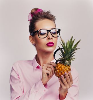 Blonde woman in glasses holding pineapple and llok at it through magnifying glass investigating quality and usefulness