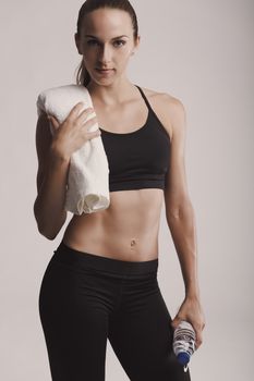 Portrait of sporty young woman holding a water bottle and towel, posing against a gray background