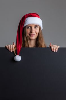 Woman wearing santa christmas hat holding and pointing blank billboard or placard sign copy space for text advertising