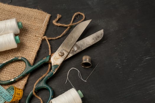 Retro sewing accessories - scissors, tape measure, thread on black wooden background. Horizontal closeup composition.