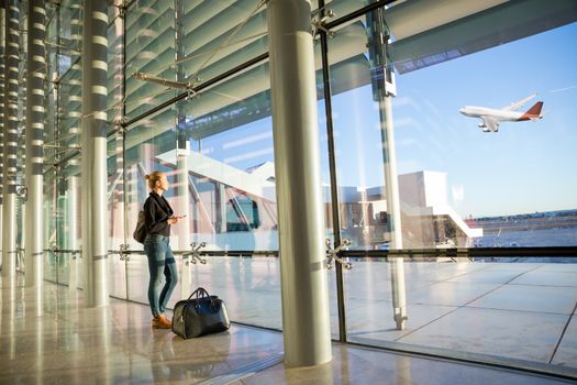 Young casual female traveler at airport, holding smart phone device, looking through the airport gate windows at planes on airport runway.