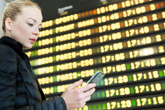 Young woman at international airport looking at the flight information board, holding smart phone in her hand, checking flight informations on phone application.