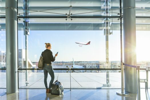 Young casual female traveler at airport, holding smart phone device, looking through the airport gate windows at planes on airport runway.