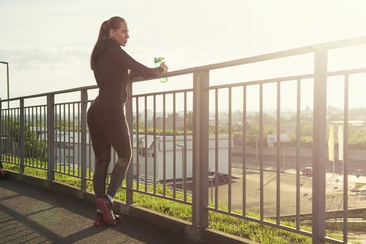 A woman in sportswear with bottle of water have rest at sunset on the evening city background