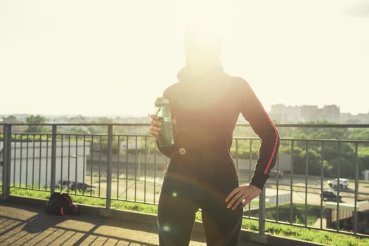 A woman in sportswear with bottle of water have rest at sunset on the evening city background