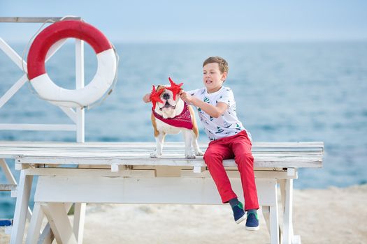 Handsome boy teen happyly spending time together with his friend bulldog on sea side Kid dog holding playing two sea stars close to life buoy float wearing red pants trousers slippers and t-shirt.