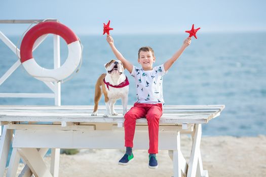 Handsome boy teen happyly spending time together with his friend bulldog on sea side Kid dog holding playing two sea stars close to life buoy float wearing red pants trousers slippers and t-shirt.