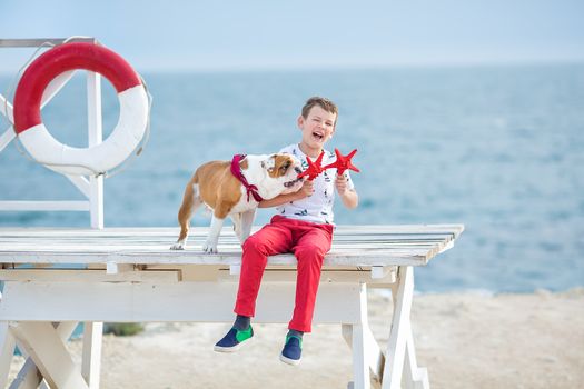 Handsome boy teen happyly spending time together with his friend bulldog on sea side Kid dog holding playing two sea stars close to life buoy float wearing red pants trousers slippers and t-shirt.