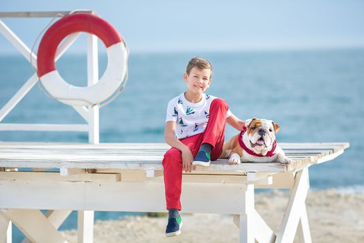 Handsome boy teen happyly spending time together with his friend bulldog on sea side Kid dog holding playing two sea stars close to life buoy float wearing red pants trousers slippers and t-shirt.