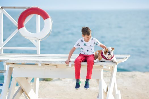 Handsome boy teen happyly spending time together with his friend bulldog on sea side Kid dog holding playing two sea stars close to life buoy float wearing red pants trousers slippers and t-shirt.