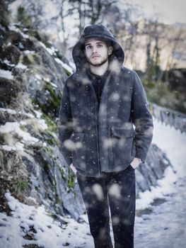 Portrait of young man in hoodie posing with hands in pockets in snowfall looking at camera.