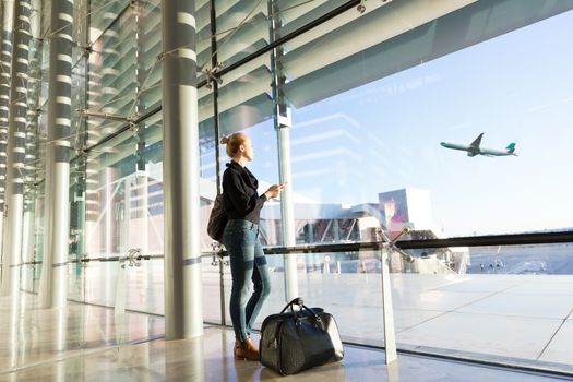 Young casual female traveler at airport, holding smart phone device, looking through the airport gate windows at planes on airport runway.