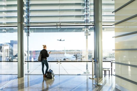 Young casual female traveler at airport, holding smart phone device, looking through the airport gate windows at planes on airport runway.