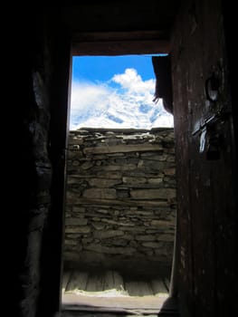 Nice view inside a small cabin through the brown wooden door, with snowy mountains in the background on a sunny day.