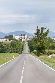 a straight road in the countryside with the village of Colloredo di Montalbano, in Italy, in the background