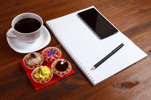 White coffee cup with notebook, four cupcakes, smartphone and pen on a wooden table.