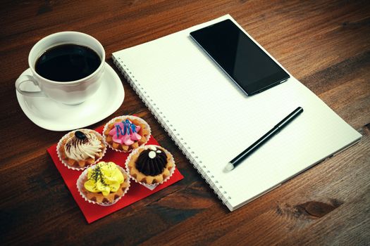 White coffee cup with notebook, four cupcakes, smartphone and pen on a wooden table.