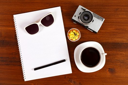 Retro film photo camera, sunglasses, cupcake, cup of coffee and notebook with pencil on brown table. Top view.