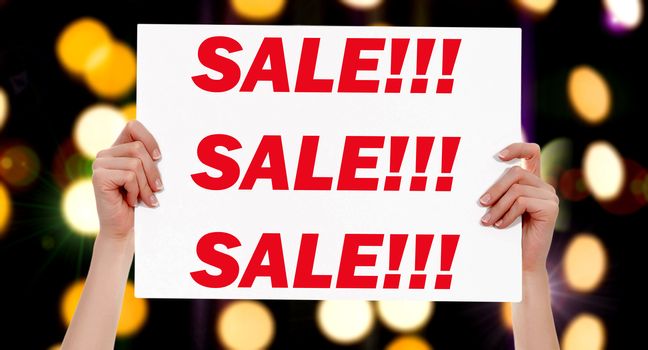 Sale! Female hands holding a placard with abstract lights bokeh background. Commercial appeal concept