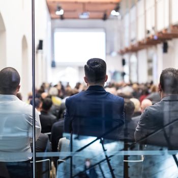 Speaker Giving a Talk at Business Meeting. Audience in the conference hall. Business and Entrepreneurship. Shot trough glass. Focus on unrecognizable woman in the audience.