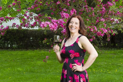 forty something brunette woman, wearing a summer dress, in front of a cherry tree, having a glass of red wine