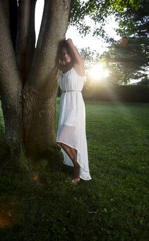 twenty something woman standing outside in the sun, wearing a white dress, leaning on a three