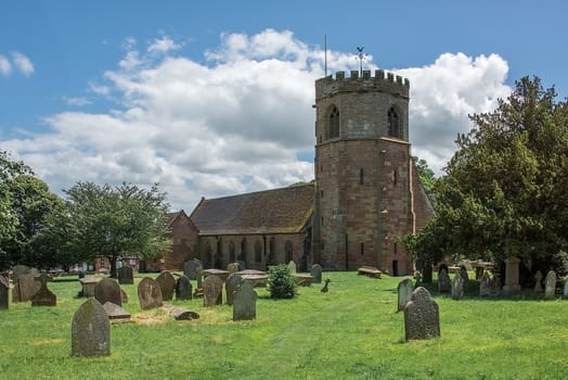 A horizontal image of a typical english country village church standing in a cemetery and taken on a sunny day with a blue sky