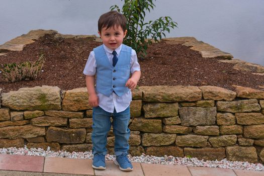 Little boy in suit with vest and tie stands in front of a wall of sand stones.