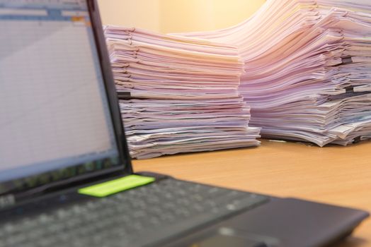 computer notebook and documents on desk stack up high waiting to be managed.