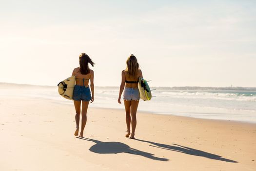 Two beautiful friends holding their surfboards and walking on the beach