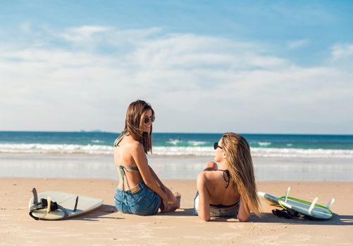 Two beautiful female friends at the beach sitting on the sand close to her surfboards while looking to the ocean
