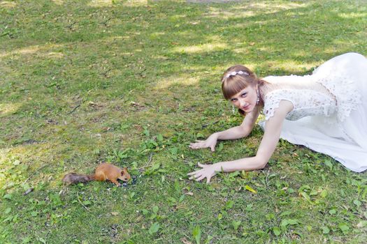 Bride in white lying on green grass next to the squirrel in summer