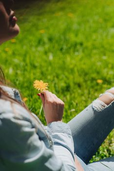 Young woman smelling dandelion flower sitting on a lawn under a tree in park