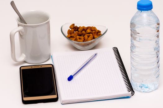 mobile phone and notebook on the desk with bowl of huzelnuts, bottle of water and the cup of coffee for keeping energy while working