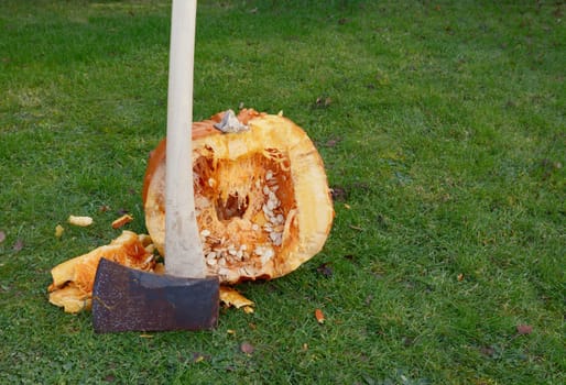 Axe stands against a large orange pumpkin, roughly cut in half outdoors, spilling flesh and seeds - with copy space