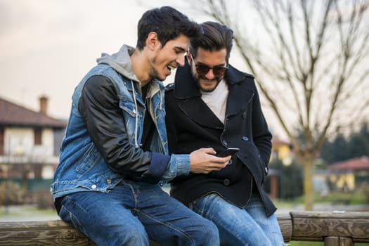 Two trendy casual young men sitting together on a bench outdoors reading an sms or text message, or looking at photos on a mobile phone