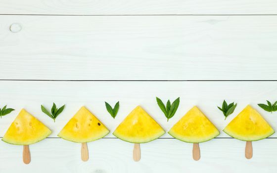 Bright yellow watermelon slices on wooden sticks on a white wooden background. Flat lay, top view, copy space