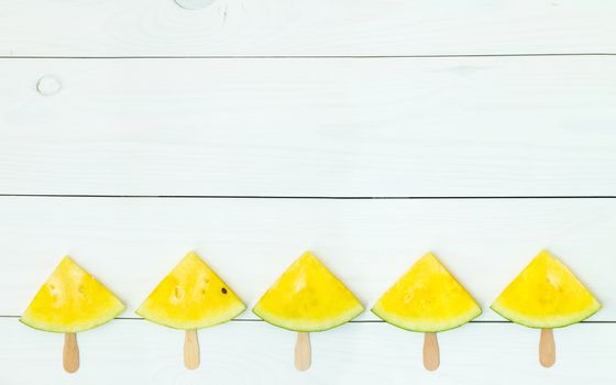 Bright yellow watermelon slices on wooden sticks on a white wooden background. Flat lay, top view, copy space